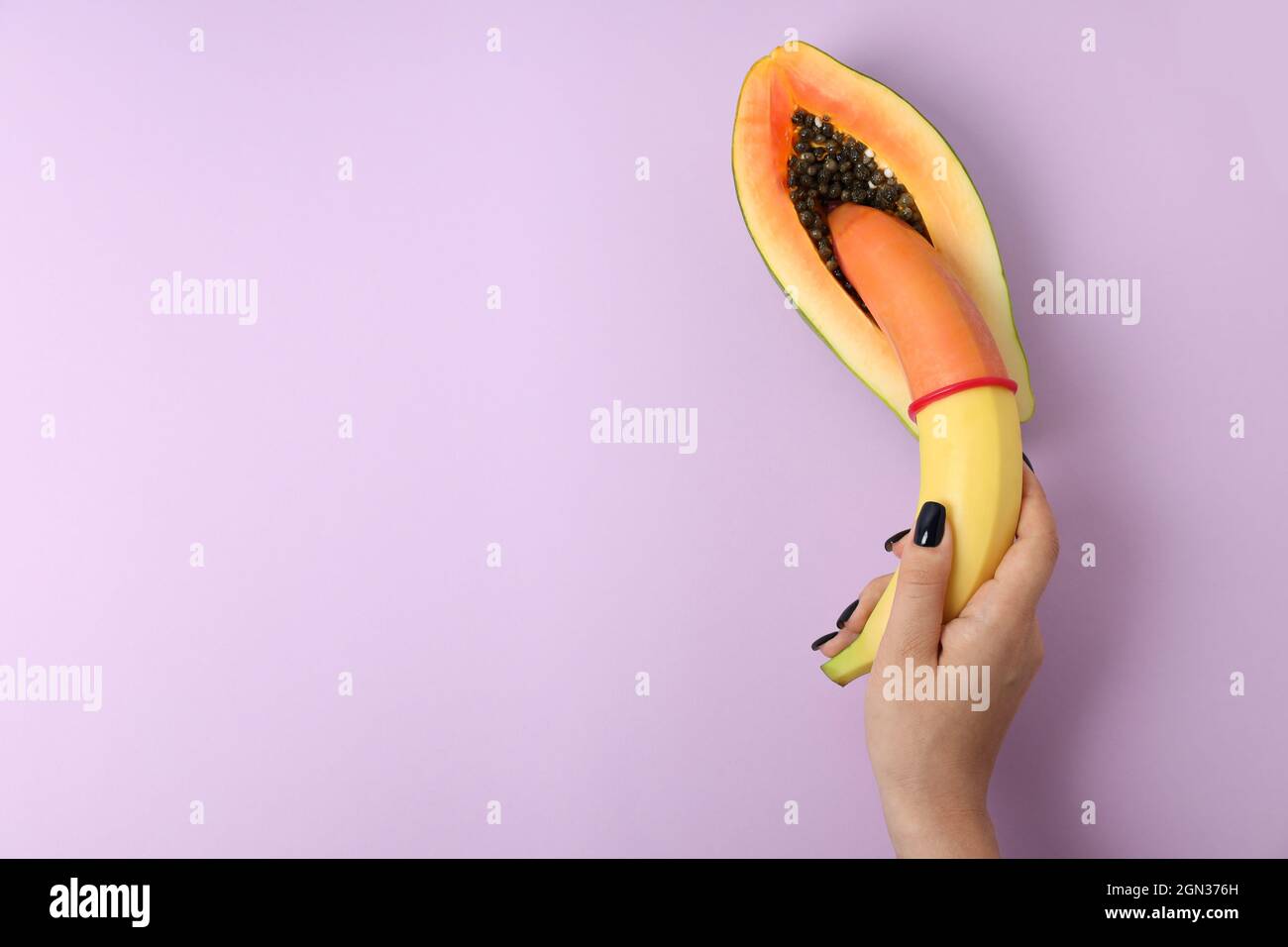 female-hand-holding-banana-with-condom-in-papaya-on-violet-background-2GN376H.jpg